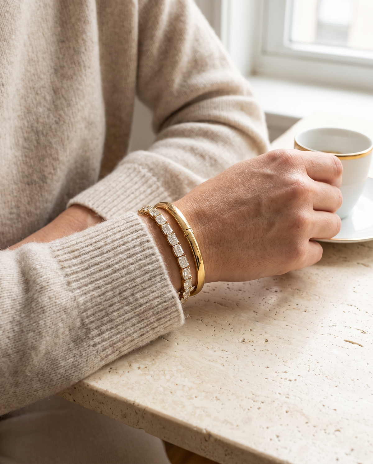 Emerald-cut diamond tennis bracelet and gold bangle on a wrist holding an espresso cup
